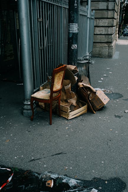 A collection of bulky waste items placed on the pavement outside a building, including a vintage upholstered chair with wooden legs and fabric covering, alongside open cardboard boxes filled with vari