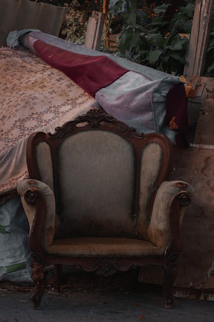 A collection of bulky waste items placed on the pavement outside a building, including a vintage upholstered chair with wooden legs and fabric covering, alongside open cardboard boxes filled with vari