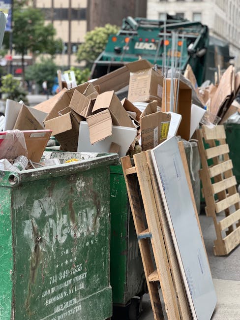 Outside a large green waste skip filled with cardboard boxes, foam packaging, and wooden pallets, ready for disposal as part of a home relocation or moving process. Next to the skip, a wooden pallet l
