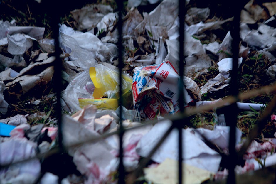 Outside a large green waste skip filled with cardboard boxes, foam packaging, and wooden pallets, ready for disposal as part of a home relocation or moving process. Next to the skip, a wooden pallet l